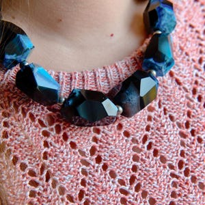 May include: A close-up of a necklace made of large, faceted, dark blue and black gemstone beads. The necklace is worn on a person wearing a pink and white knitted sweater.