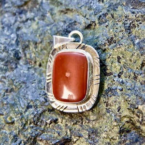 May include: A silver pendant featuring a rectangular, reddish-brown stone. The stone is set in a silver frame with a decorative border. The pendant has a loop at the top for a chain. The background is a textured, grey-green stone.