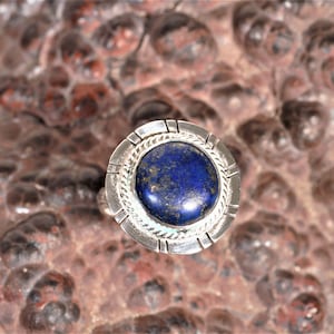 May include: A silver ring featuring a deep blue lapis lazuli stone set in a circular bezel. The ring has a decorative border around the stone.
