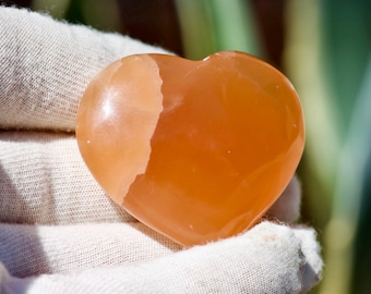 May include: Orange heart-shaped stone, possibly a crystal or gemstone, with a smooth, polished surface. The stone is held in a white fabric. The heart shape is clearly defined, with a lighter, almost white, section on one side.