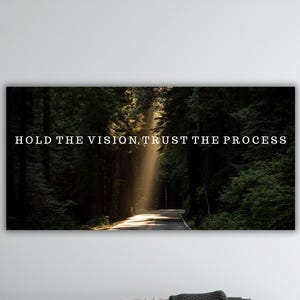 May include: A rectangular wall art print featuring a forest scene with a road leading into the distance. The text "HOLD THE VISION, TRUST THE PROCESS" is displayed in white across the top of the image. The image is framed and mounted on a white wall.