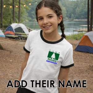 May include: A white t-shirt with black trim around the neck and sleeves. The shirt features a green and blue camp logo with the text "CAMP" and "NAME". The bottom of the image reads "ADD THEIR NAME".