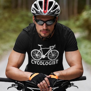 May include: A black t-shirt with a white bicycle graphic and the word "CYCOLOGIST" below it. The person in the image is wearing a white, red, and black bicycle helmet, black sunglasses, and black and orange cycling gloves.