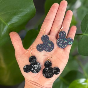 以下が含まれることがあります： ゴールドカラーのフックが付いた、ミッキーマウス型のイヤリング4組。2組はシルバーの斑点のある黒色で、他の2組はカラフルな斑点のある濃い青色です。イヤリングは緑の葉を背景に展示されています。