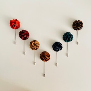May include: Seven different colored fabric flower lapel pins. The pins are arranged in a V shape on a white background. The colors are red, burgundy, brown, tan, dark blue, and green.