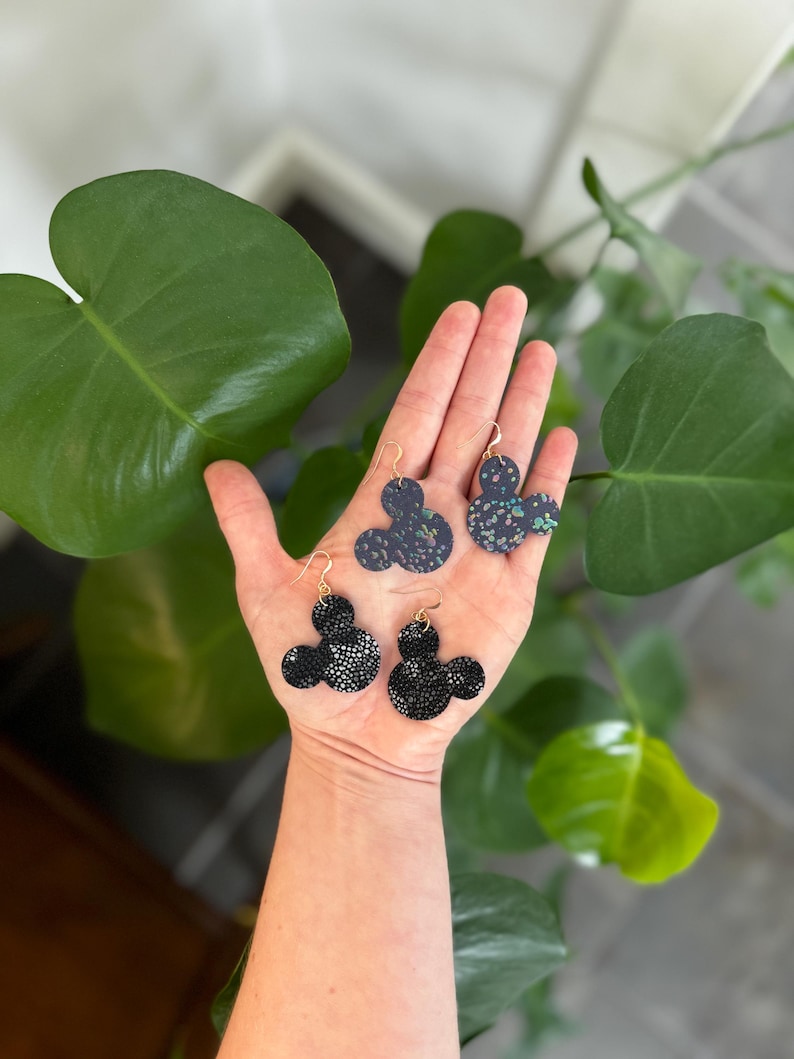 以下が含まれることがあります： ゴールドカラーのフックが付いた、ミッキーマウス型のイヤリング4組。2組はシルバーの斑点のある黒色で、他の2組はカラフルな斑点のある濃い青色です。イヤリングは緑の葉を背景に展示されています。