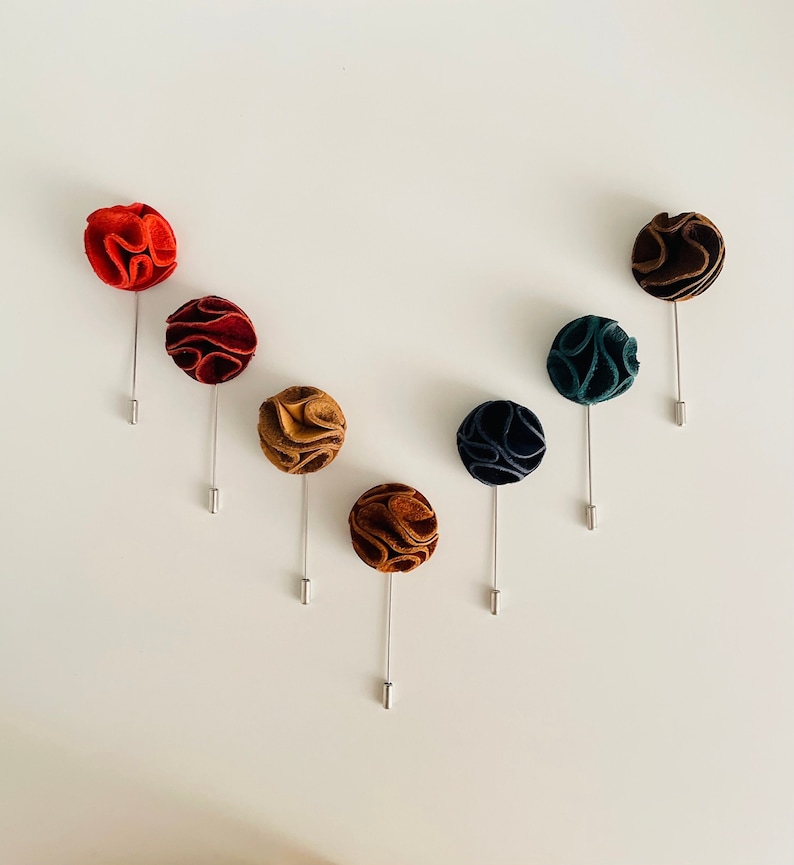 May include: Seven different colored fabric flower lapel pins. The pins are arranged in a V shape on a white background. The colors are red, burgundy, brown, tan, dark blue, and green.