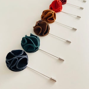 May include: A set of seven fabric flower lapel pins in various colors, including brown, red, teal, and black. Each pin features a small, round flower with a silver pin.