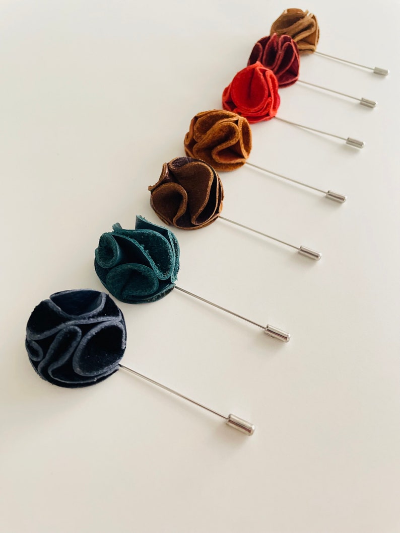 May include: A set of seven fabric flower lapel pins in various colors, including brown, red, teal, and black. Each pin features a small, round flower with a silver pin.