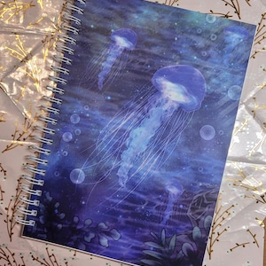 May include: Spiral-bound notebook featuring a vibrant illustration of jellyfish in shades of blue and purple. The cover art depicts several jellyfish with long, flowing tentacles, set against a deep blue ocean background with bubbles and aquatic plants.