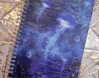 May include: Spiral-bound notebook featuring a vibrant illustration of jellyfish in shades of blue and purple. The cover art depicts several jellyfish with long, flowing tentacles, set against a deep blue ocean background with bubbles and aquatic plants.