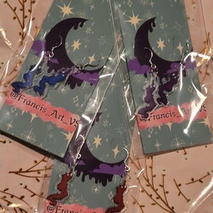 May include: Three pairs of dangle earrings with a crescent moon, clouds, and dripping paint design. The earrings are made of clear acrylic and have silver hooks. The earrings are packaged on cards with a starry night background and the text "@Francis_Art_VS".