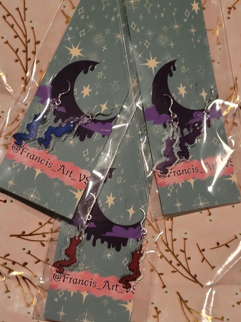 May include: Three pairs of dangle earrings with a crescent moon, clouds, and dripping paint design. The earrings are made of clear acrylic and have silver hooks. The earrings are packaged on cards with a starry night background and the text "@Francis_Art_VS".