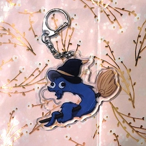May include: A whimsical blue creature keychain with a witch hat and broom. The keychain has a silver metal clasp and chain. The background is a pink floral pattern.