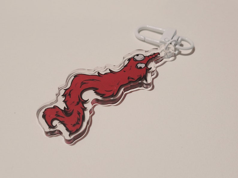 May include: A red worm-like creature with two eyes is printed on a clear acrylic keychain. The keychain has a white metal ring and clasp.