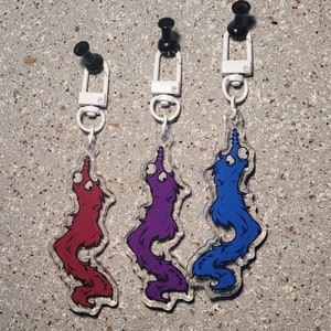 以下が含まれることがあります： 紐に付いたミミズのキーホルダーが3つ。ミミズは赤、紫、青。それぞれのミミズは、飛び出た目で、白い紐がついています。