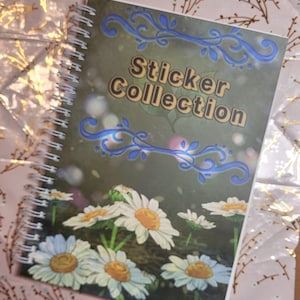 May include: A spiral-bound notebook titled "Sticker Collection" with a cover featuring white daisies with yellow centers against a green background. Blue decorative swirls frame the title. The notebook rests on a pink and gold patterned surface.