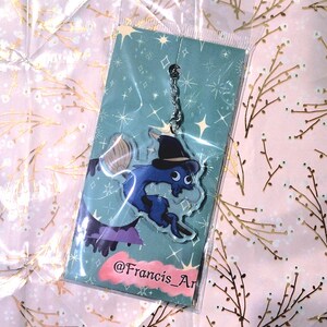 May include: A blue and black acrylic charm featuring a cartoon creature wearing a witch hat, with a broom and purple accents. The charm is on a teal background with stars and is packaged in a clear plastic sleeve. The text "@Francis_Ar" is visible.