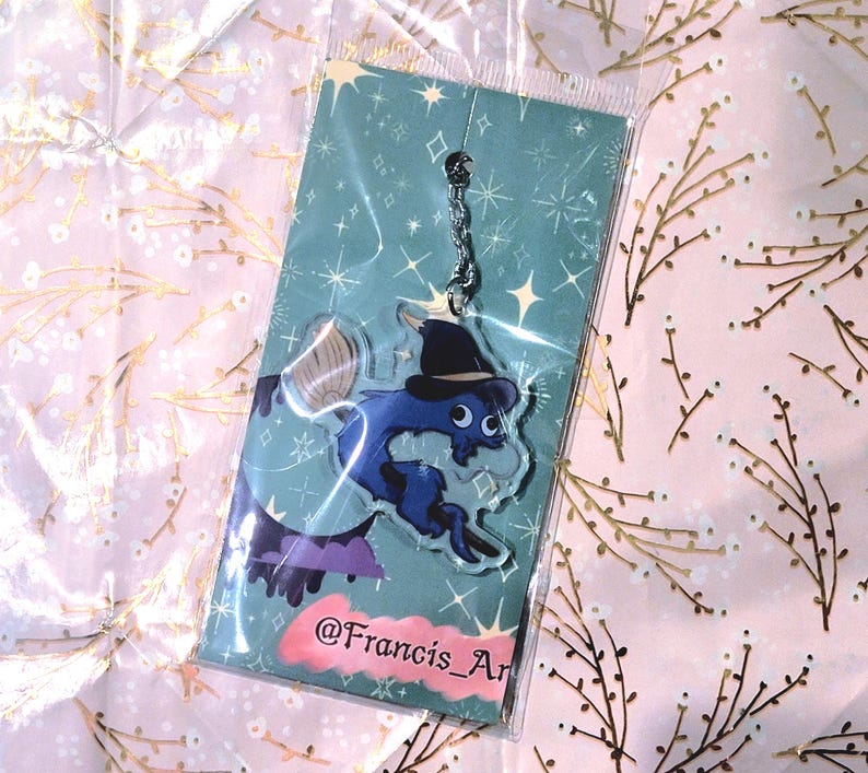 May include: A blue and black acrylic charm featuring a cartoon creature wearing a witch hat, with a broom and purple accents. The charm is on a teal background with stars and is packaged in a clear plastic sleeve. The text "@Francis_Ar" is visible.
