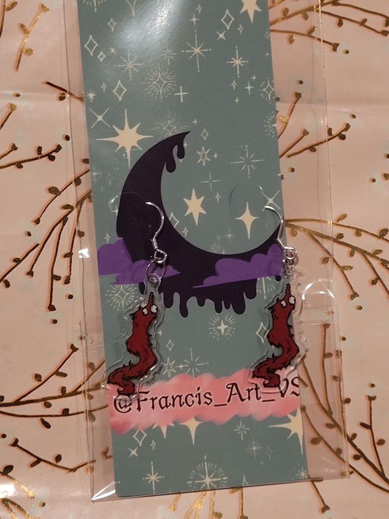 May include: A pair of red worm earrings with a dripping black moon and stars on a blue background. The earrings are on a card with the text "@Francis_Art_VS".