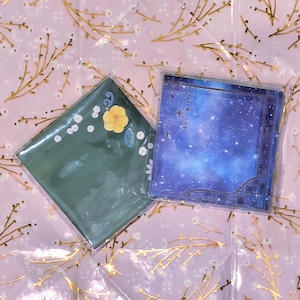 May include: Two square decorative paper sheets. One is green with a yellow flower and white accents. The other is blue with a starry night design and a decorative border. Both are in clear plastic packaging.