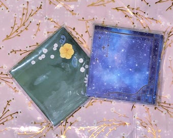 Könnte beinhalten: Zwei quadratische dekorative Papierbögen. Einer ist grün mit einer gelben Blume und weißen Akzenten. Der andere ist blau mit einem Sternennacht-Design und einem dekorativen Rand. Beide sind in transparenter Kunststoffverpackung.