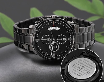 Op de afbeelding: Zwart chronograafhorloge met een zwarte metalen band. De wijzerplaat van het horloge heeft een zwarte achtergrond met witte markeringen en een datumscherm. De achterkant van het horloge heeft een gegraveerd bericht met de tekst "You're that "Once in a lifetime" dream come true. I LOVE YOU every second of my LIFE."