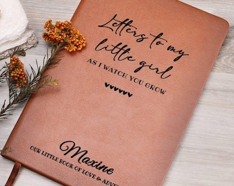 May include: A brown leather journal with the text "Letters to my little girl" and "Maxine" in black script. The journal has a ribbon bookmark and is decorated with a sprig of orange flowers.