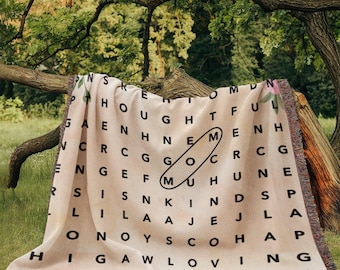 May include: A beige woven throw blanket with a black word search pattern. The pattern includes the words "thoughtful", "kind", "loving", and "go", among others. The blanket has fringe on all sides.