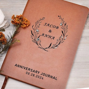 May include: A brown leather-bound journal with the text "Anniversary Journal 08.28.2024" and a floral wreath design with the names "Jacob & Anna" in the center.