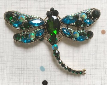 May include: A gold-tone dragonfly brooch with green and blue rhinestones. The dragonfly's wings are spread out and its body is adorned with a large green rhinestone.