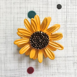 May include: A yellow metal sunflower brooch with a brown center. The brooch is on a white background with black and green polka dots.