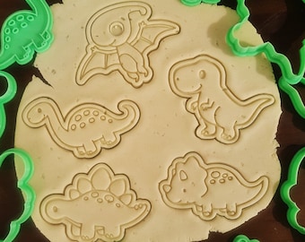 Pode incluir: Um conjunto de 11 cortadores de biscoitos de plástico verde em forma de dinossauros. Os cortadores estão dispostos sobre uma superfície de madeira marrom.