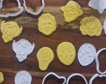 May include: A collection of 14 white and yellow plastic cookie cutters shaped like horror movie characters, including Jason Voorhees, Freddy Krueger, and Pennywise the Clown. The cutters are arranged on a wooden surface.