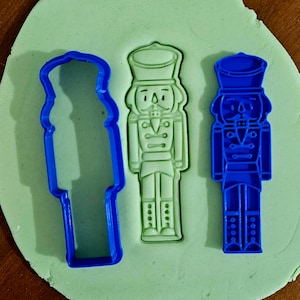 May include: Three blue plastic cookie cutters shaped like nutcrackers. The cutters are on a green surface.