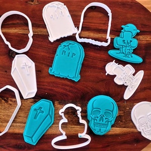 May include: A set of 10 Halloween-themed cookie cutters. The cutters are made of white and teal plastic and feature various spooky designs, including skulls, tombstones, and coffins. The cutters are arranged on a wooden surface.