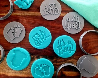 May include: A set of nine grey and teal silicone cookie cutters. The cutters are shaped like a baby onesie, a pair of footprints, and the phrases "It's a Girl!" and "It's a Boy!"
