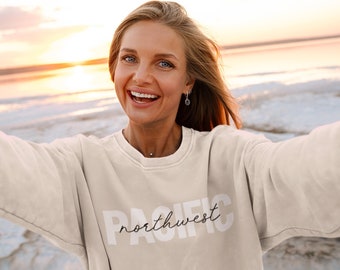 May include: A woman wearing a light beige sweatshirt with the text "PACIFIC northwest" printed in white on the front. She is smiling and standing in front of a body of water with a sunset in the background.