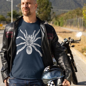 May include: A navy blue t-shirt with a white graphic of a spider. The person wearing the shirt is also wearing a black leather jacket and blue jeans.