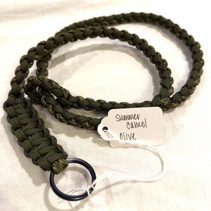 May include: Olive green lanyard made of braided cord, featuring a black metal ring. A white tag attached reads "Summer Camo Olive". The lanyard is designed for carrying keys or an ID badge.