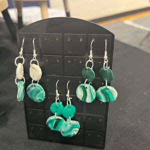 May include: Three pairs of dangle earrings with green and white clay beads. The earrings are displayed on a black gridded earring display card.