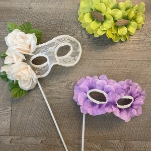 Puede incluir: Tres máscaras de carnaval en palos. Las máscaras están hechas de flores de tela. Una máscara es blanca con encaje, otra es verde y la última es morada.