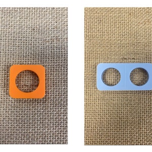 May include: Two plastic components: an orange square with a circular cutout and a light blue rectangular piece with two circular cutouts. The items are displayed on a textured, tan surface.