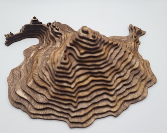May include: A wooden model of a mountain range with layered contours, creating a 3D effect. The model is made of light brown wood with a natural grain pattern.