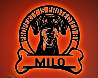 May include: A black metal sign with the words "PROPERTY IS PROTECTED BY" in a circle around a dog's head, with the name "MILO" below. The sign is illuminated with an orange glow, and the text "NAME CAN BE CHANGED" is in a green box.