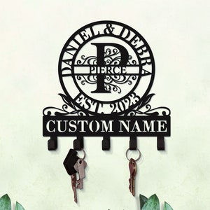 May include: Black metal key holder with a circular design featuring the names "Daniel & Debra" and the initial "P" with the last name "Pierce" and the text "Est. 2023". The key holder has five hooks and the text "Custom Name" is displayed below the circular design.