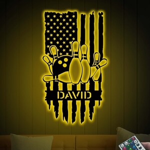 May include: A black and yellow night light with a distressed American flag design, bowling pins and a bowling ball, and the name "DAVID" in the center. The light is illuminated and has a remote control.