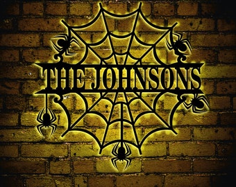 Puede incluir: Una silueta de metal negro de una telaraña con arañas colgando de la telaraña. La telaraña tiene una placa de identificación que dice "The Johnsons". La silueta está iluminada con una luz amarilla cálida y montada en una pared de ladrillo.