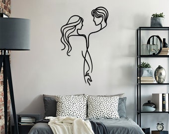 May include: Black metal wall art depicting a couple in a single line drawing. The couple is facing each other and their arms are intertwined. The art is hanging on a white wall above a gray bed with white bedding.