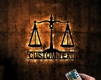 May include: A black metal silhouette of a scale of justice with the words "CUSTOM TEXT" underneath. The scale is illuminated with warm white lights and is mounted on a textured wall. A remote control with color changing buttons is visible in the bottom right corner.